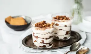 Zwei köstliche Schichten-Desserts im Glas, garniert mit Kakaopulver und Krümeln, stehen auf einem Serviertablett. Daneben liegen Löffel und eine Schale mit Keksen.