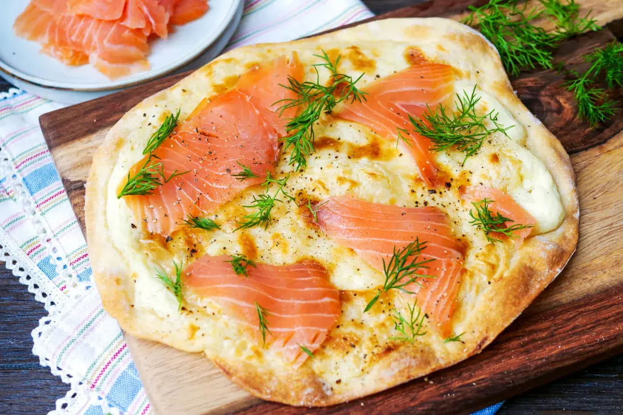 Flammkuchen mit Lachs, belegt mit zarten Lachsscheiben, cremiger Käsesauce und frischem Dill, garniert mit schwarzem Pfeffer.