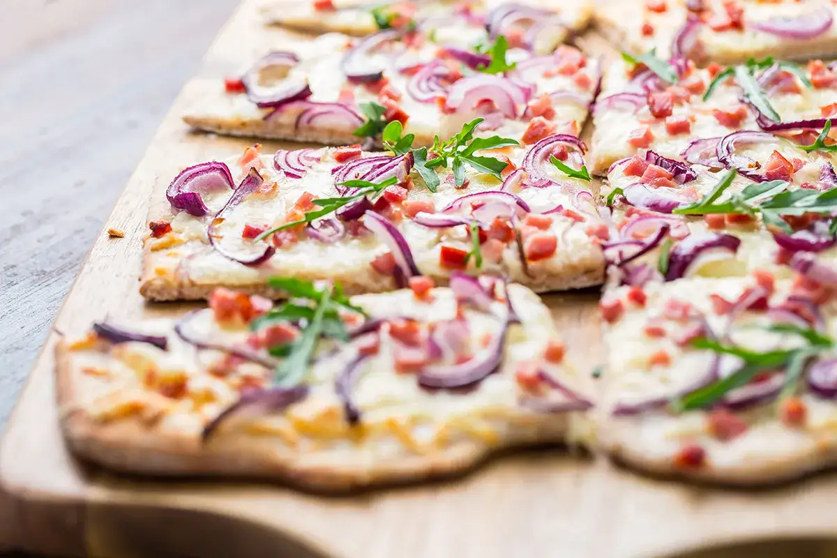 Flammkuchen-Rezept: Dünner Teig mit Käse, roten Zwiebeln und Speck belegt, garniert mit Rucola.