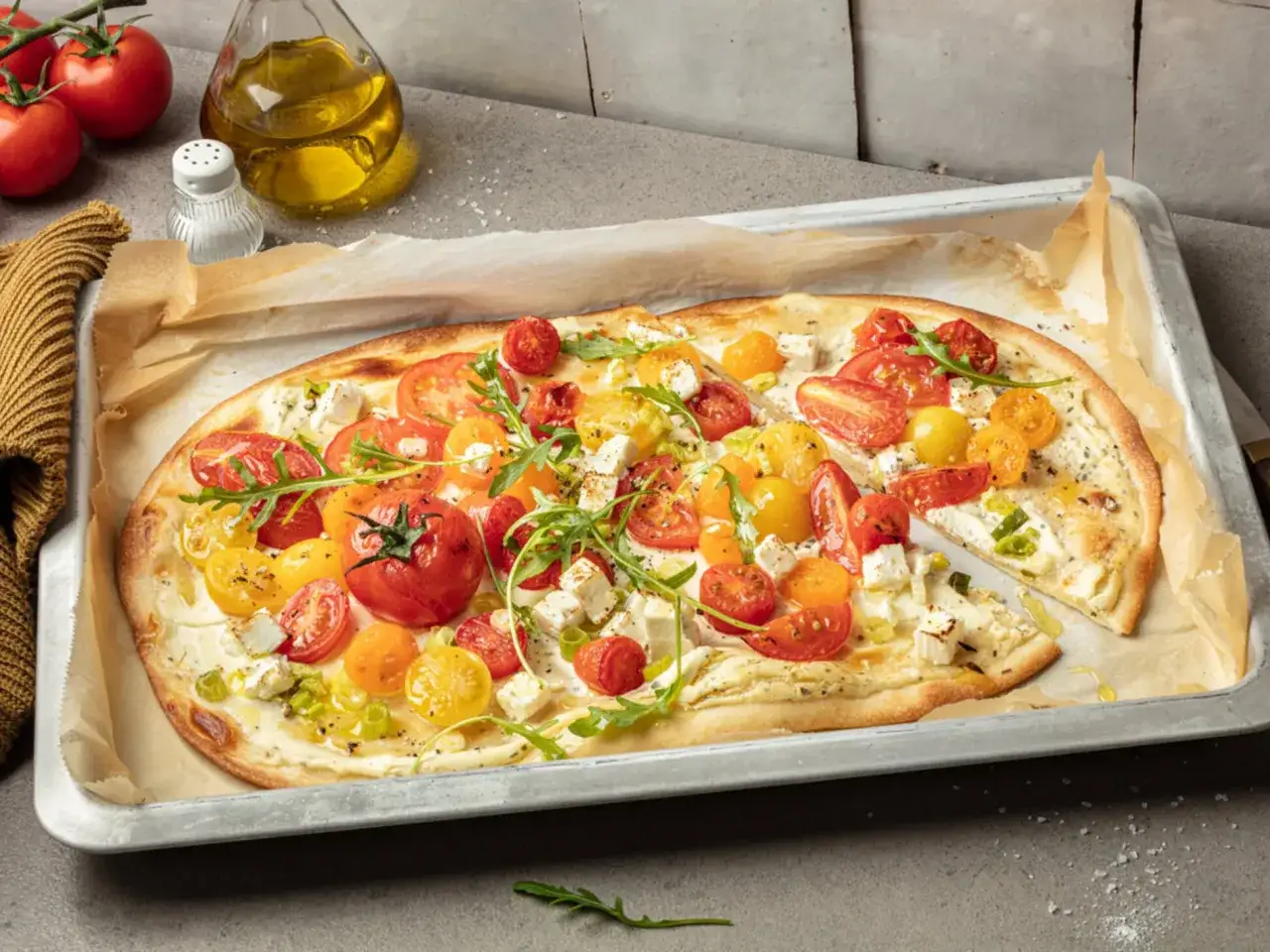 Flammkuchen vegetarisch mit bunten Tomaten, Feta und Rucola auf einem Backblech.