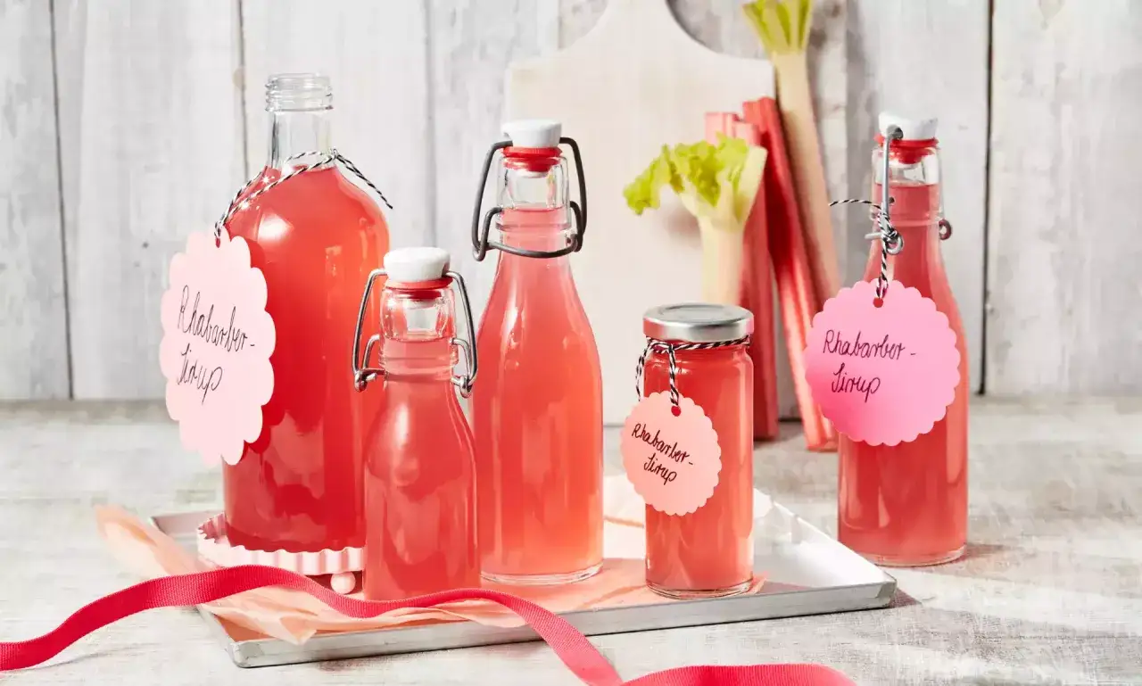 Flaschen mit selbstgemachtem Rhabarbersirup, bereit für das Rhabarbersirup Rezept. Frischer Rhabarber im Hintergrund.