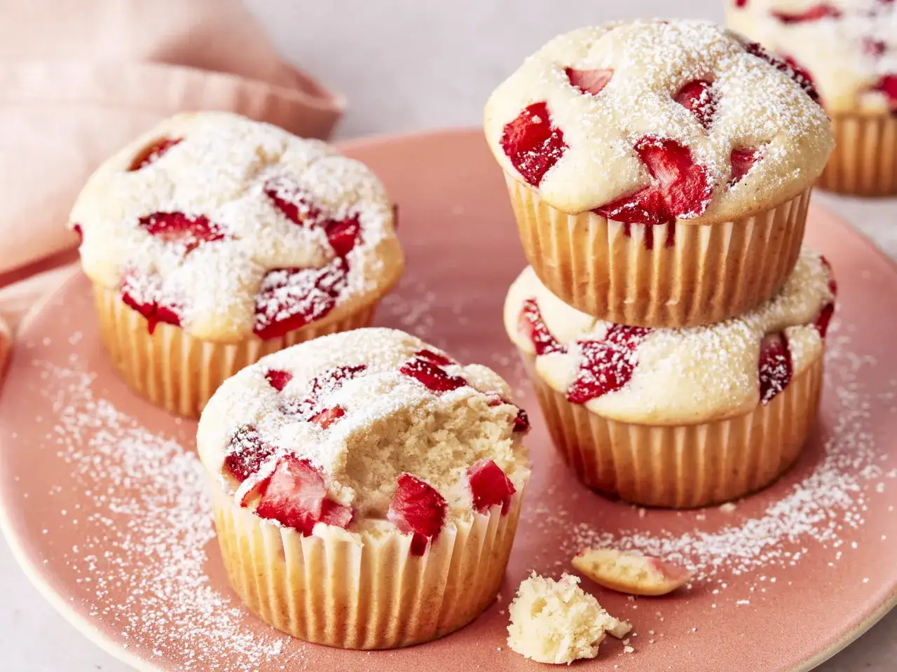 Erdbeermuffins mit Puderzucker bestäubt auf einem rosa Teller, ein Muffin ist angeknabbert.
