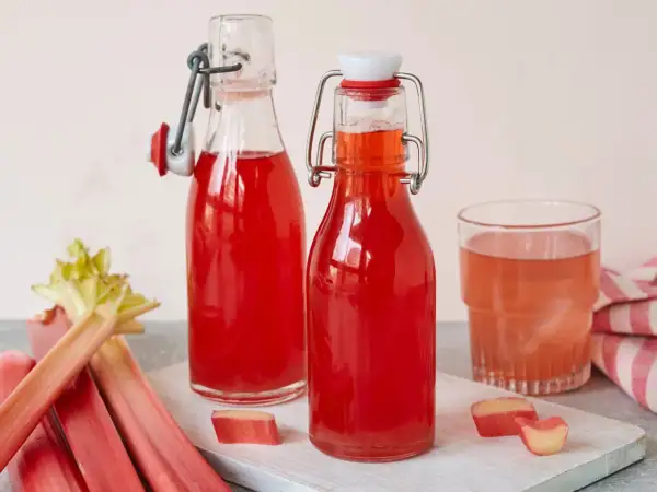 Zwei Flaschen mit leuchtend rotem Rhabarbersirup, ein Glas und frische Rhabarberstangen. Perfekt für ein erfrischendes Rhabarbersirup Rezept.