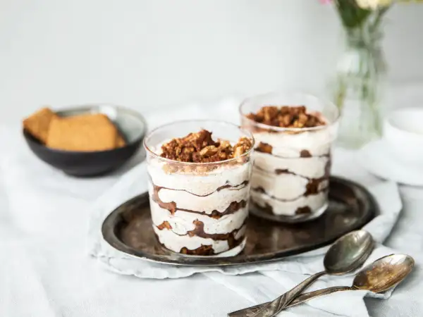 Zwei köstliche Schichten-Desserts im Glas, garniert mit Kakaopulver und Krümeln, stehen auf einem Serviertablett. Daneben liegen Löffel und eine Schale mit Keksen.