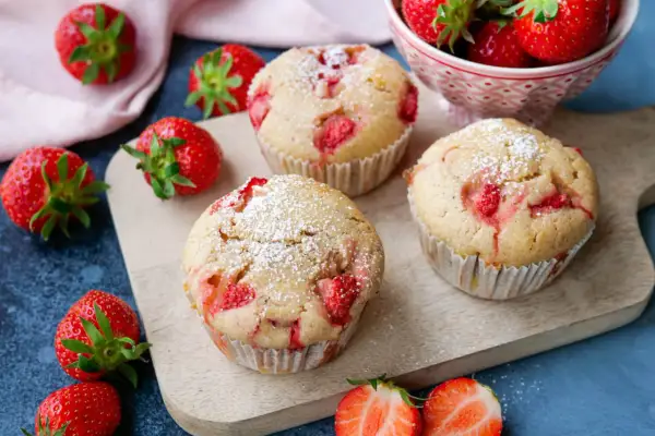 Saftige erdbeer muffins, bestäubt mit Puderzucker, liegen auf einem Holzbrett. Frische Erdbeeren umgeben die Leckereien.