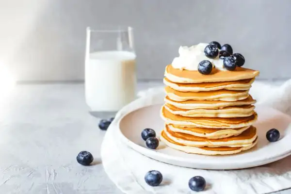 Ein Stapel goldbrauner Protein Pfannkuchen, garniert mit Sahne und Blaubeeren, daneben ein Glas Milch.