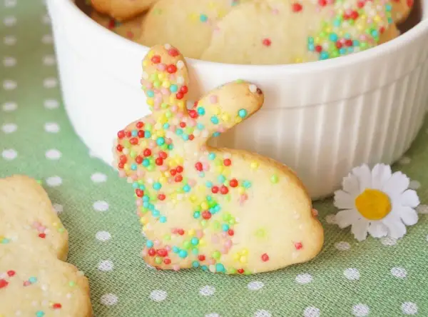 Bunte Osterkekse zum Ausstechen, verziert mit bunten Streuseln, liegen auf einem grünen Tupfentuch. Ein süßer Osterhase steht im Vordergrund.