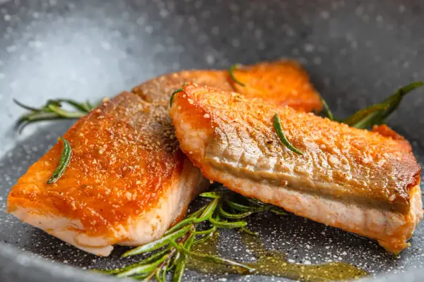 Zwei perfekt gebratene Lachsfilets mit knuspriger Haut und Rosmarin in einer Pfanne.